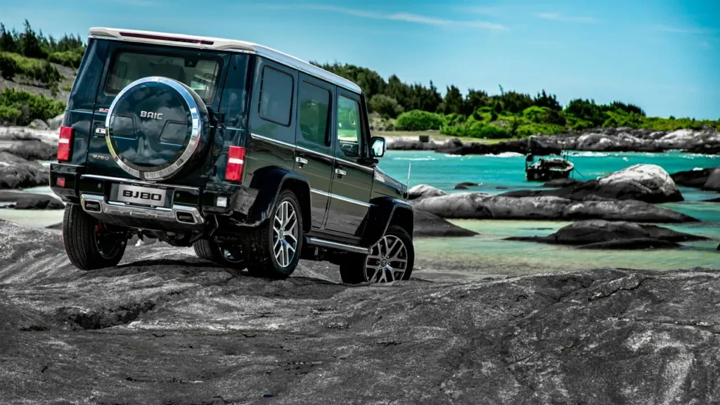 Rear three-quarter view of the BAIC BJ80 SUV parked near turquoise water and rocky shore under a blue sky.